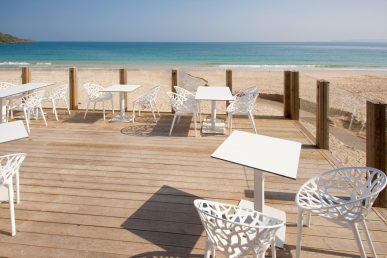 Tables and chairs on wooden decking with beach behind.